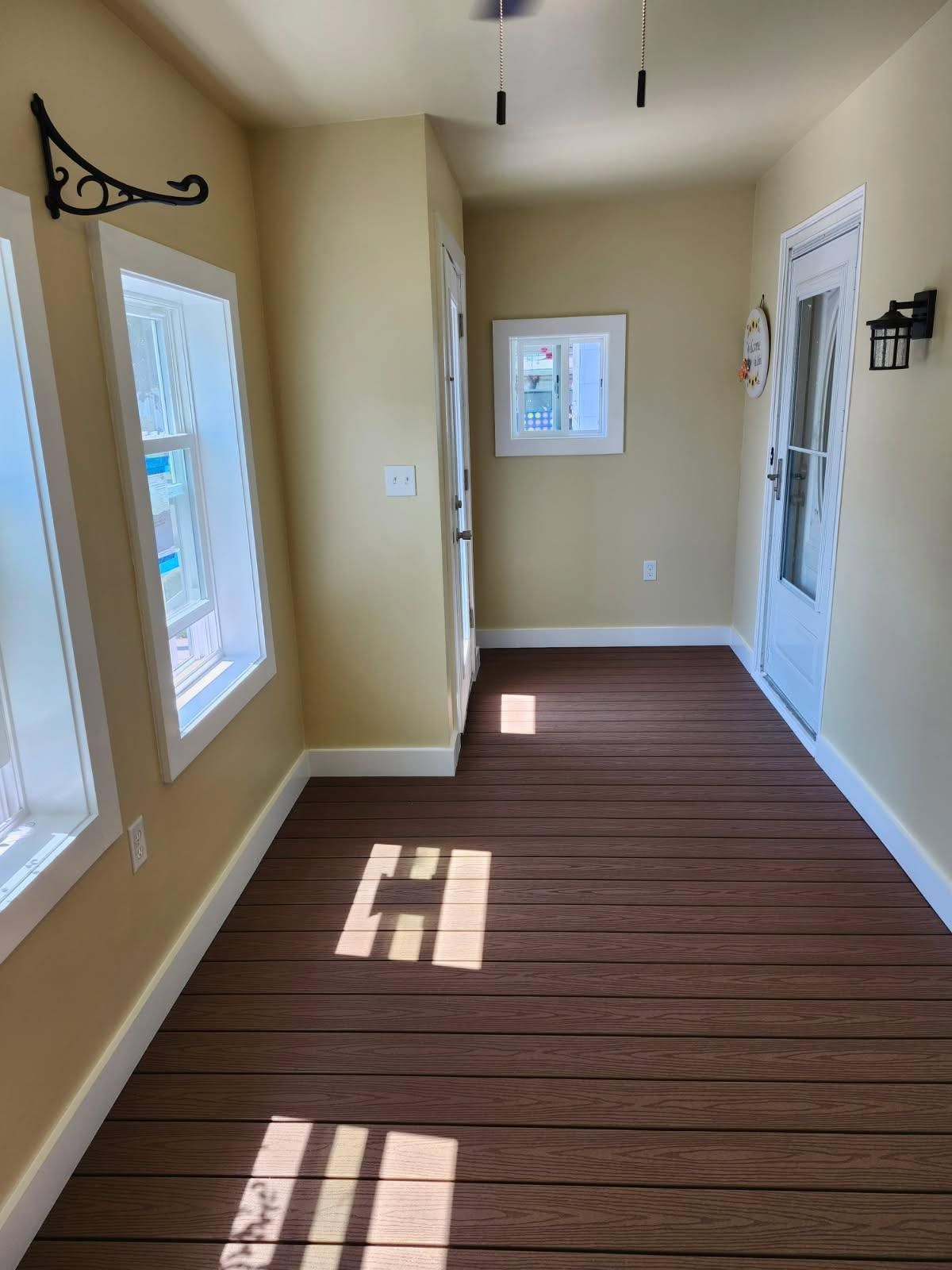 Narrow beige hallway with wood plank floor, windows, and a white door, lit by sunlight.