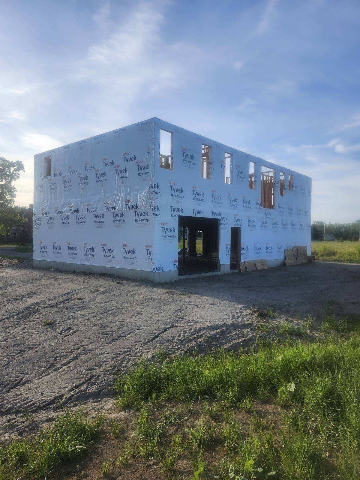 Partially built white house framed in construction wrap on a gravel lot under a blue sky