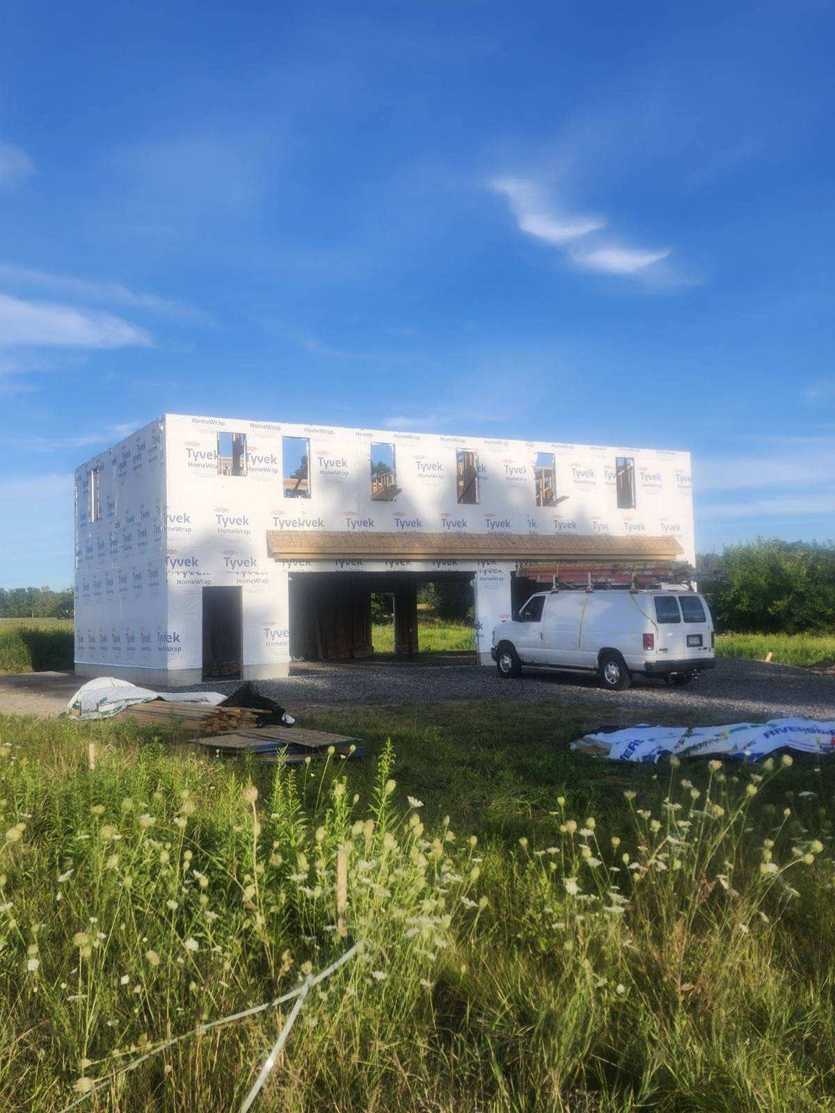 Partially built white house frame in a grassy field, with a white van parked beside it under a blue sky