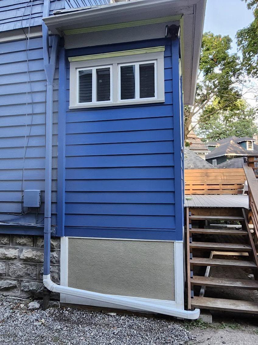 Blue house exterior with a small window, stone foundation, and wooden stairs on the right