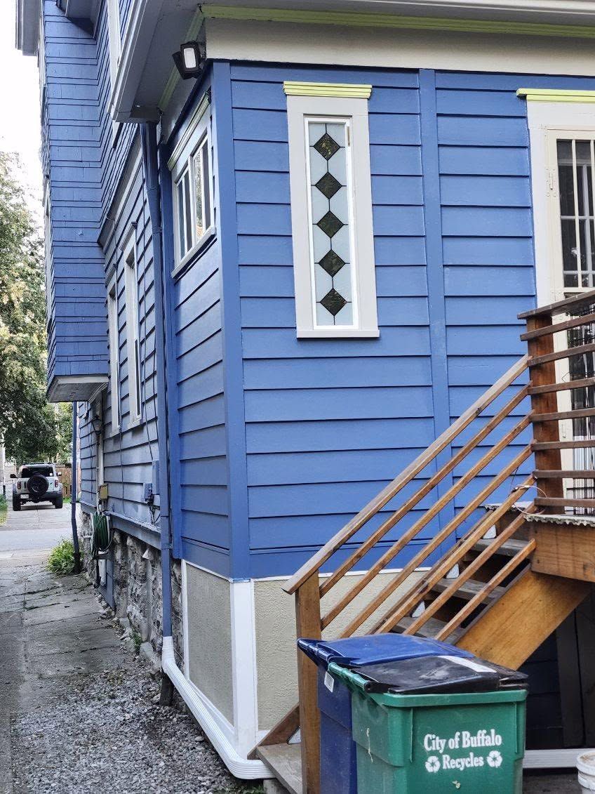 Blue house exterior with white trim, narrow side window, and wooden stairs beside a gravel alley.
