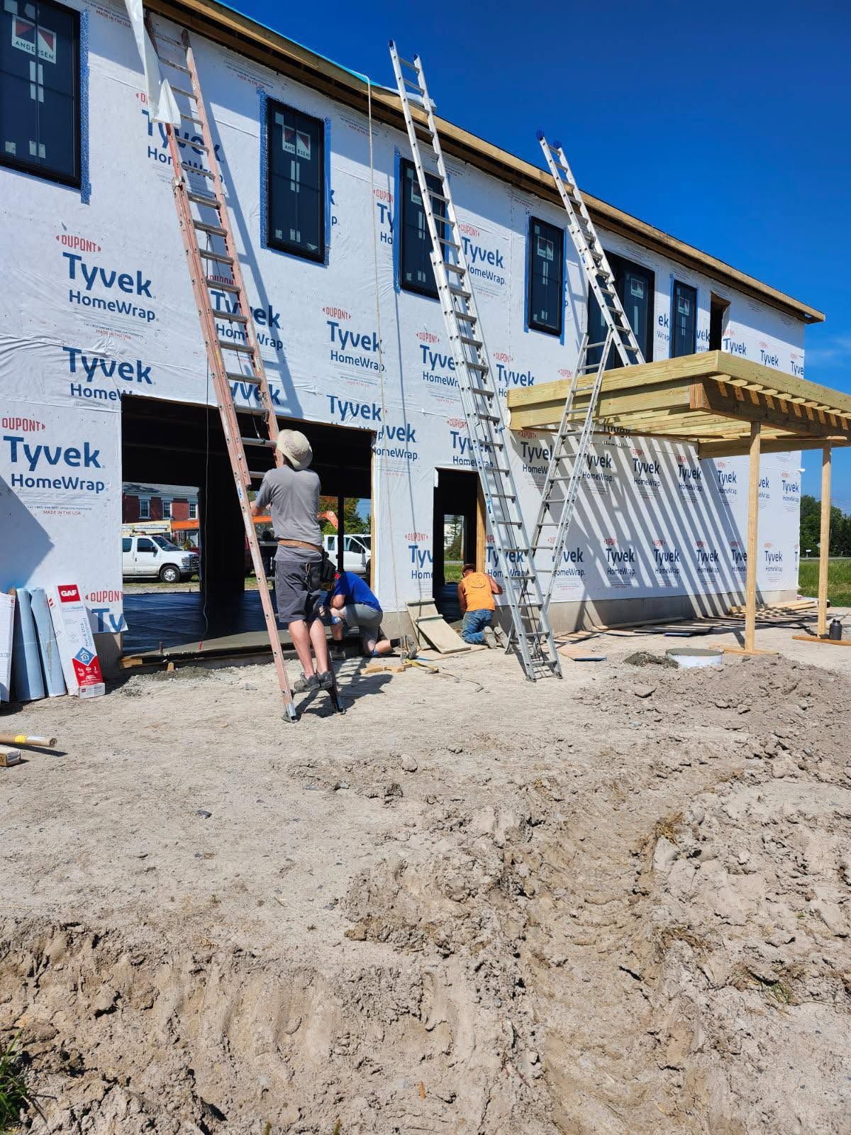 Two workers on scaffolding at a Tyvek-wrapped house under construction, with ladders and dirt foreground