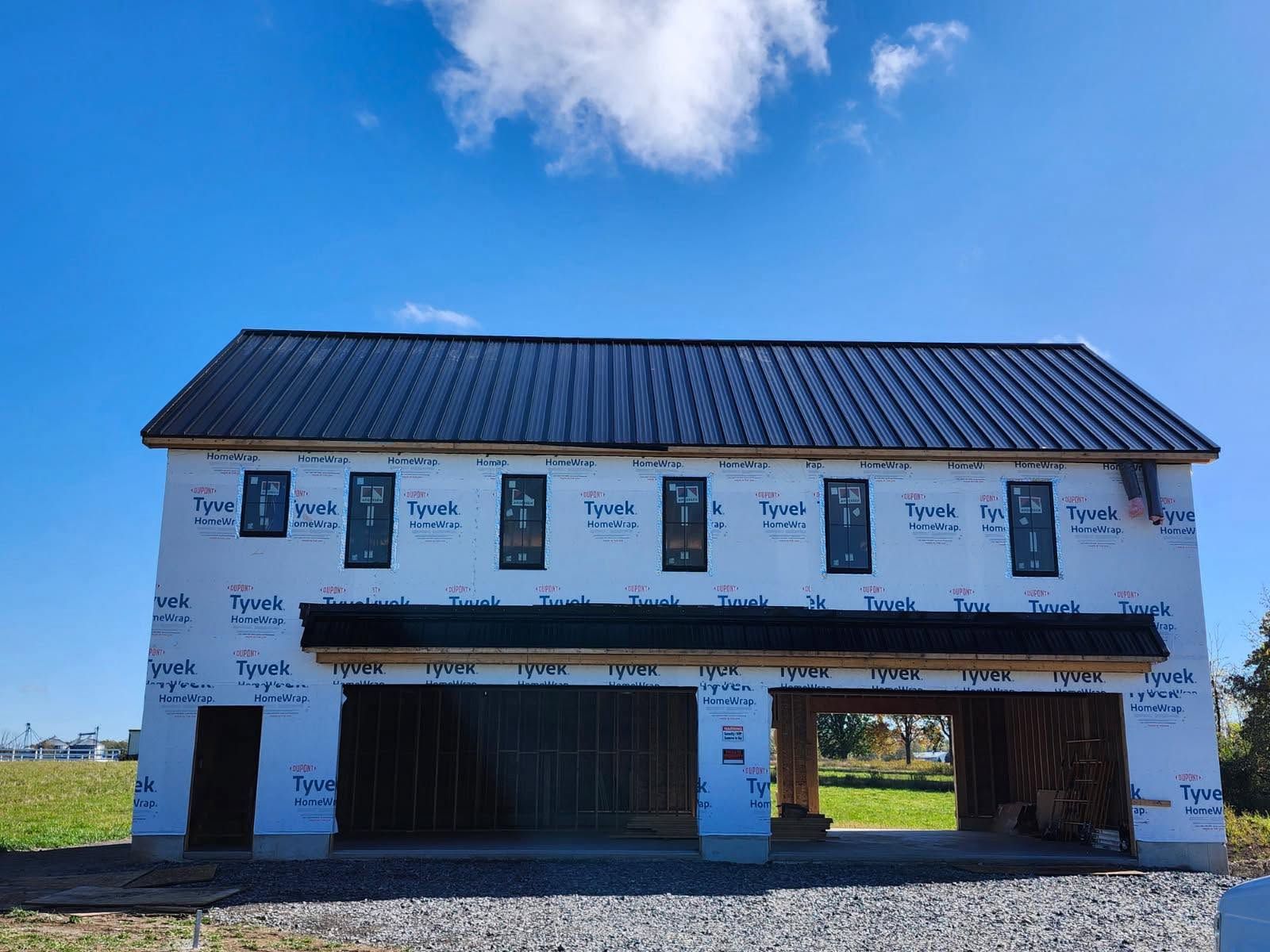 Two-story unfinished white building with a dark roof under a blue sky, set in an open field.