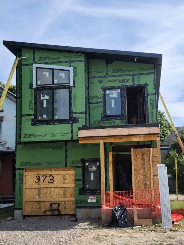 Green two-story house under construction with exposed framing, black windows, and wooden entry porch.