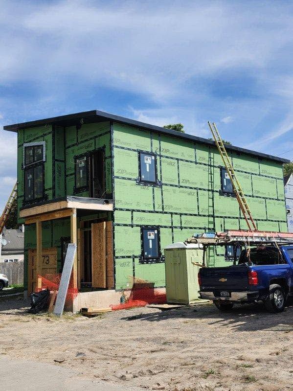 Two-story green house under construction with a ladder, truck, and clear blue sky.