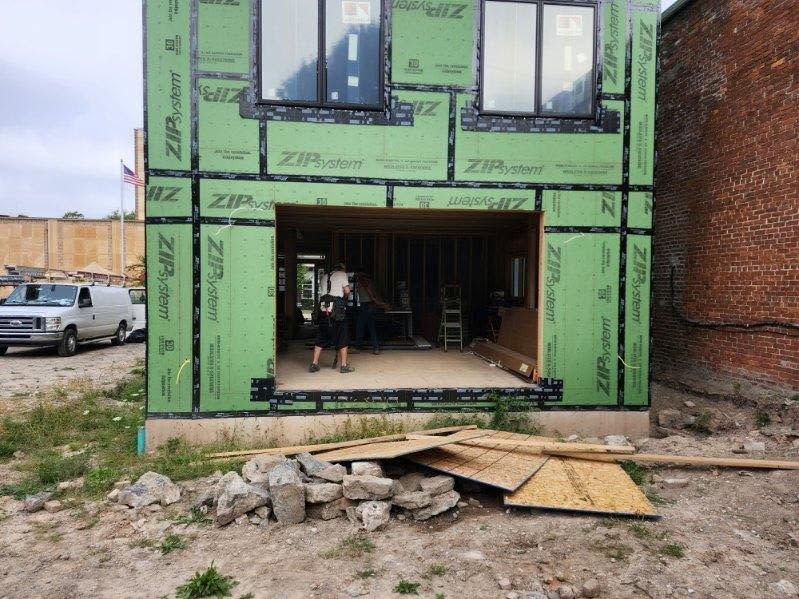 Green-framed house under construction with open garage; two people stand inside amid building materials.