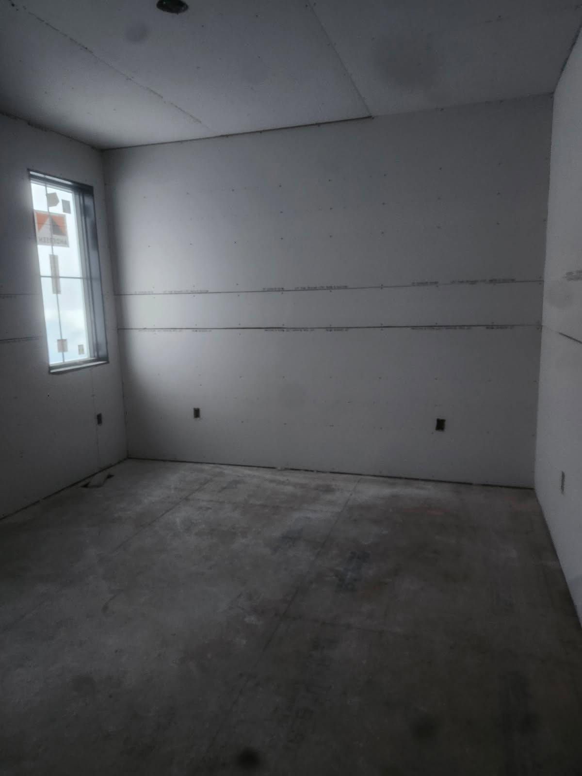 Empty unfinished room with gray concrete floor, white walls, and a small window on the left.