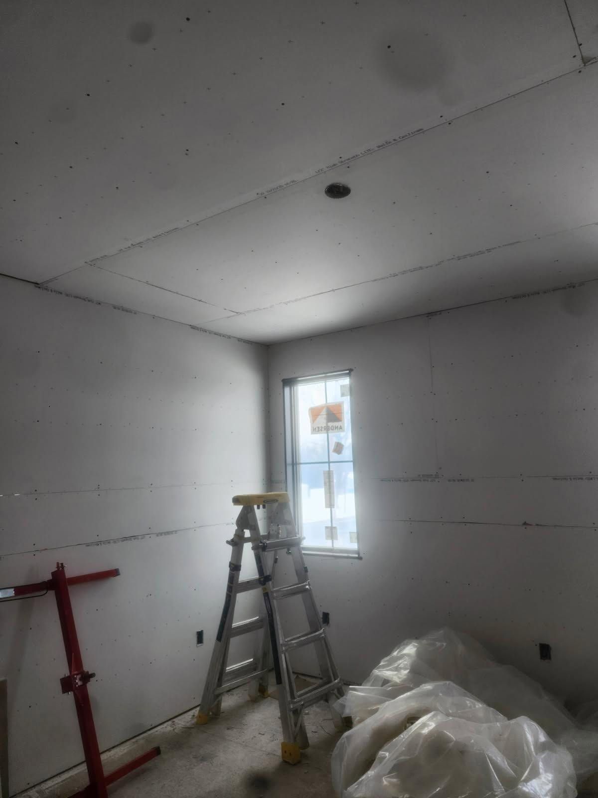 Unfinished room with drywall ceiling, ladder, red stand, and plastic-covered materials by a bright window.