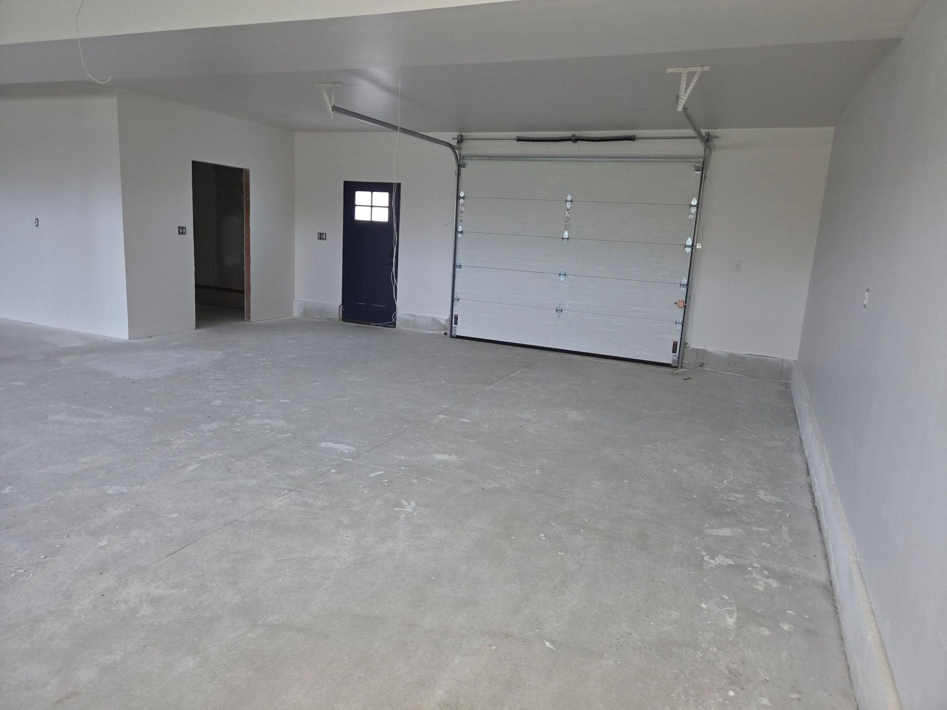 Empty unfinished garage with concrete floor, white walls, a blue door, and a white roll-up garage door.
