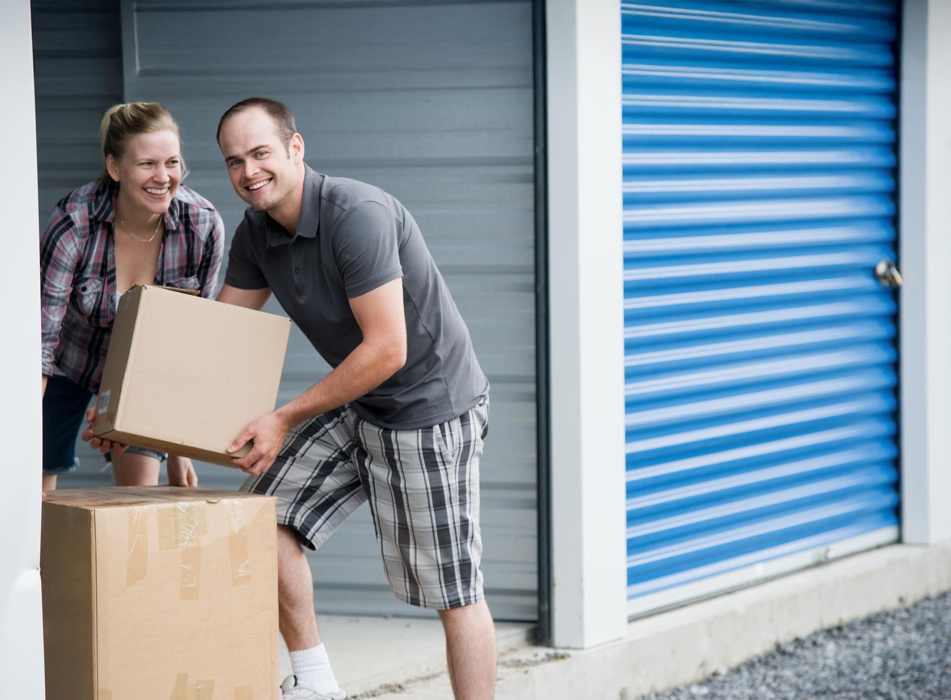 Two people carry boxes outside a storage unit with a blue roll-up door on a clear day.