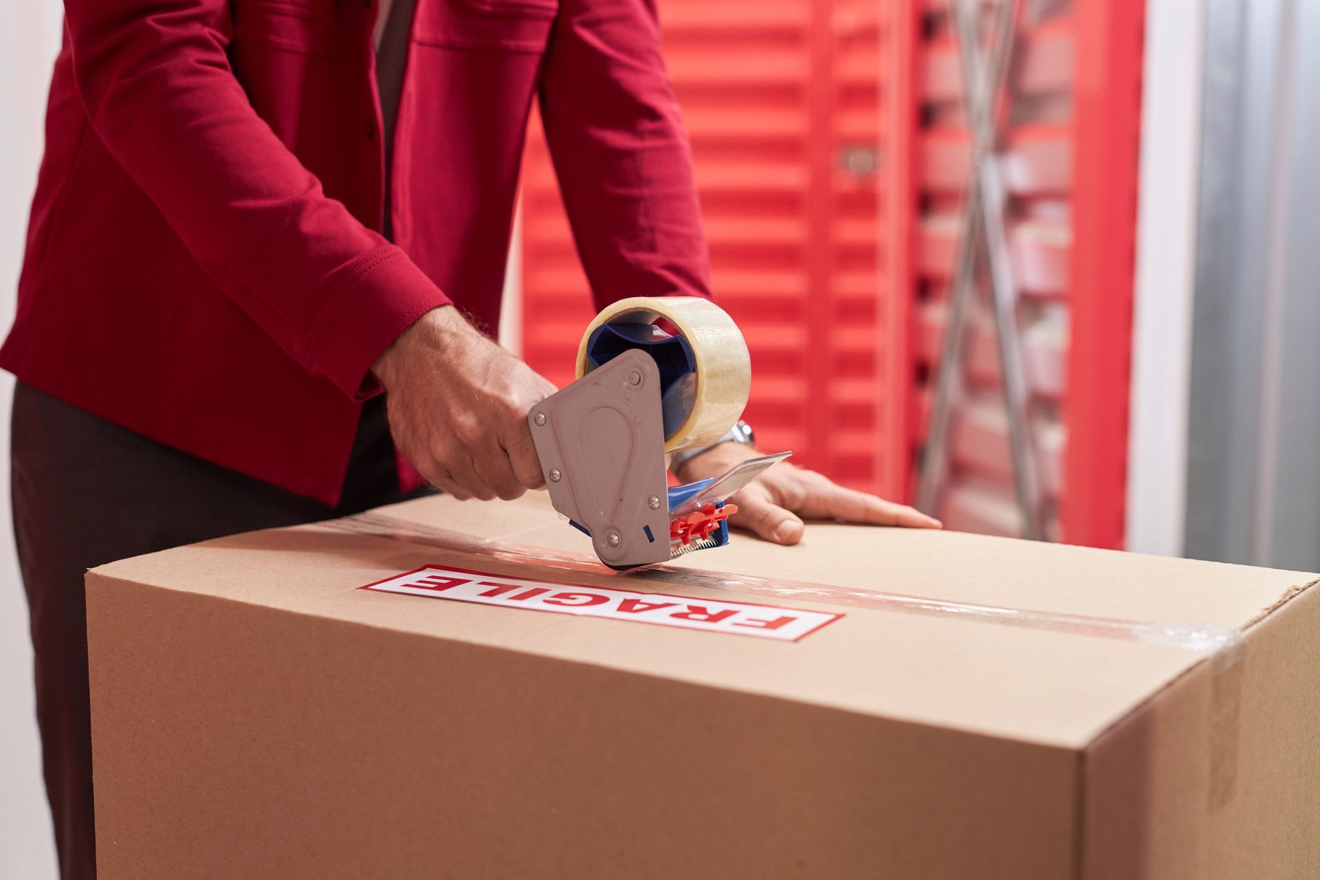 Person in a red blazer using a tape gun to seal a cardboard box marked fragile near storage units.