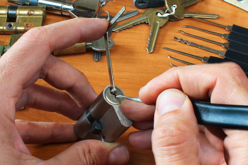 Hands using lock picks to open a silver padlock on a workbench with tools nearby