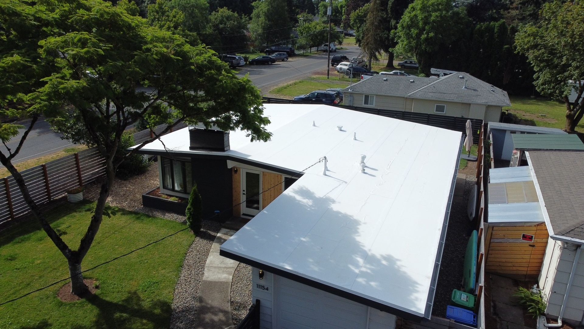 Aerial view of a one-story house with a white roof, surrounded by green lawn and trees.