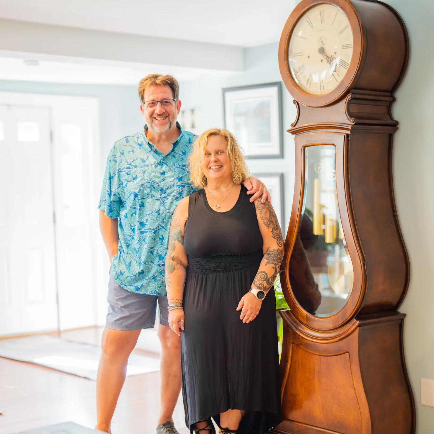 Two people standing beside a large wooden grandfather clock in a bright room