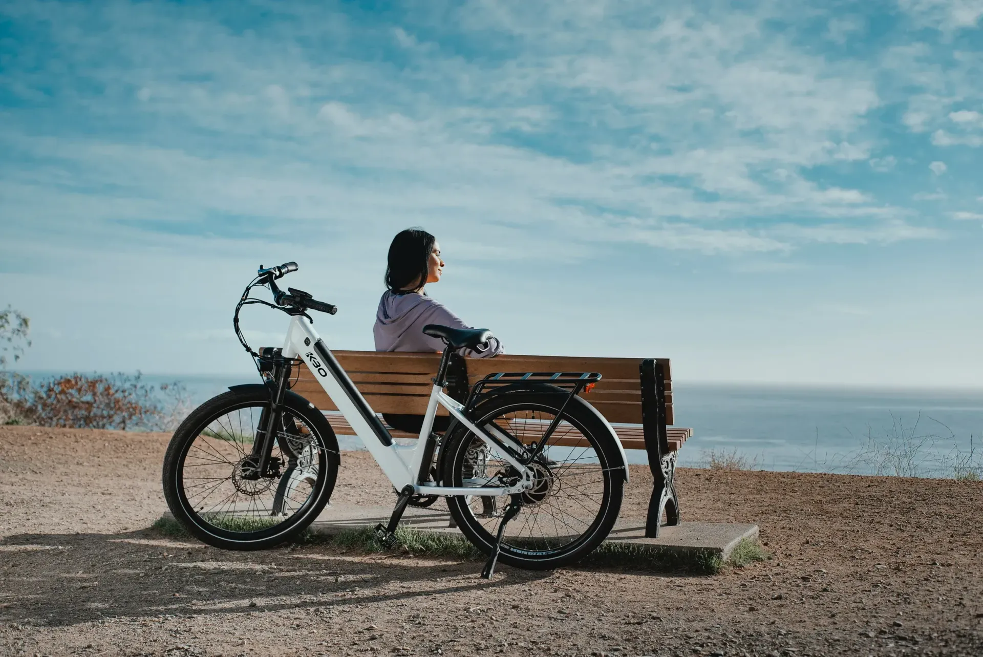 Person on a bicycle beside a wooden bench by the sea under a blue sky