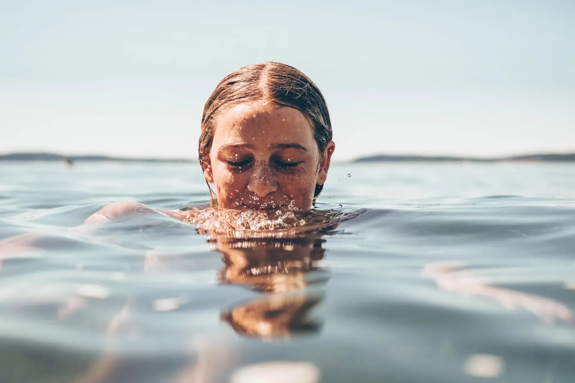Person floating face-down in calm water, eyes closed, with sunlight reflecting on the surface.