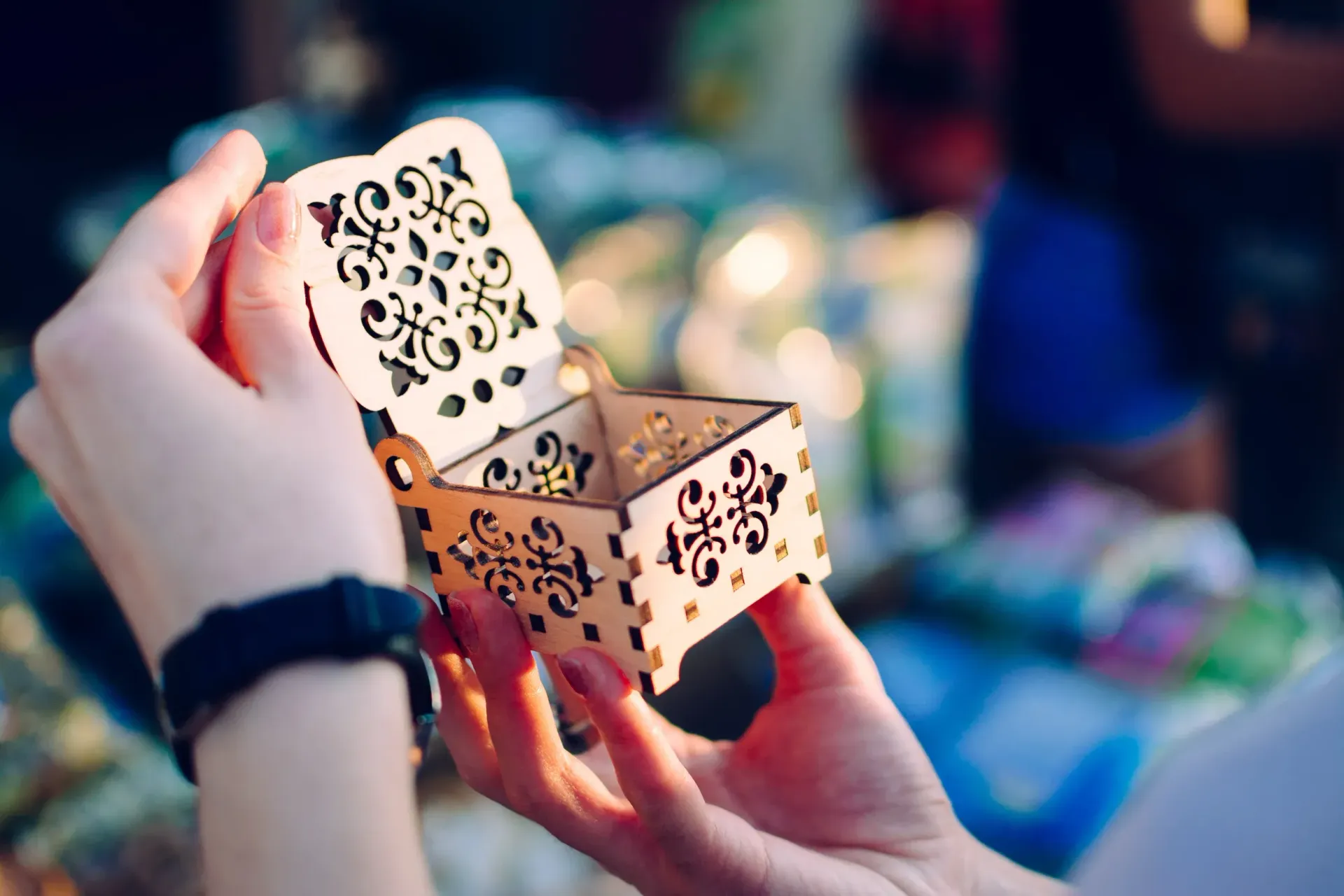 Hands holding an open decorative wooden box with floral cutouts in a blurred indoor setting