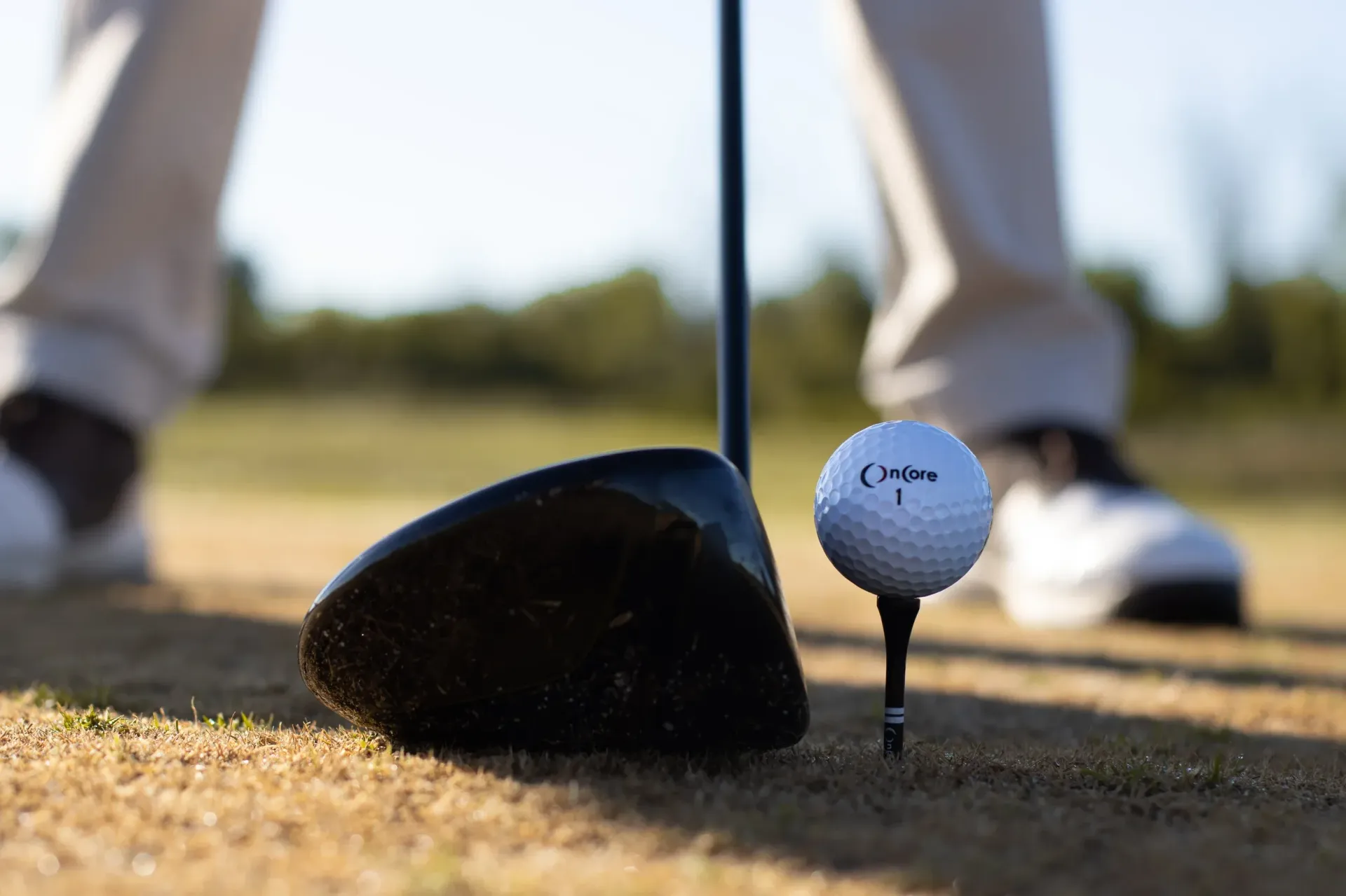 Golf ball on a tee beside a driver on a tee box, with golfer’s shoes in the background