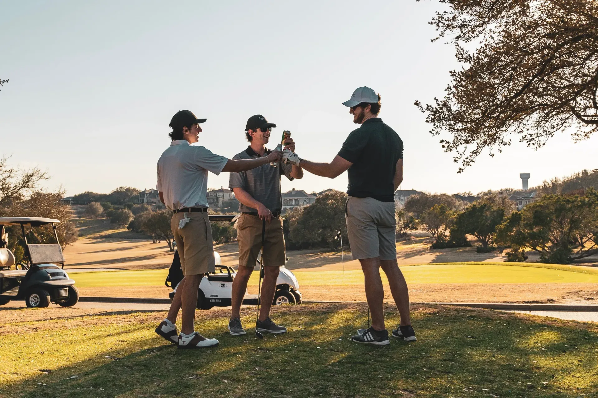 Three golfers standing on a fairway, talking and holding up drinks at sunset.