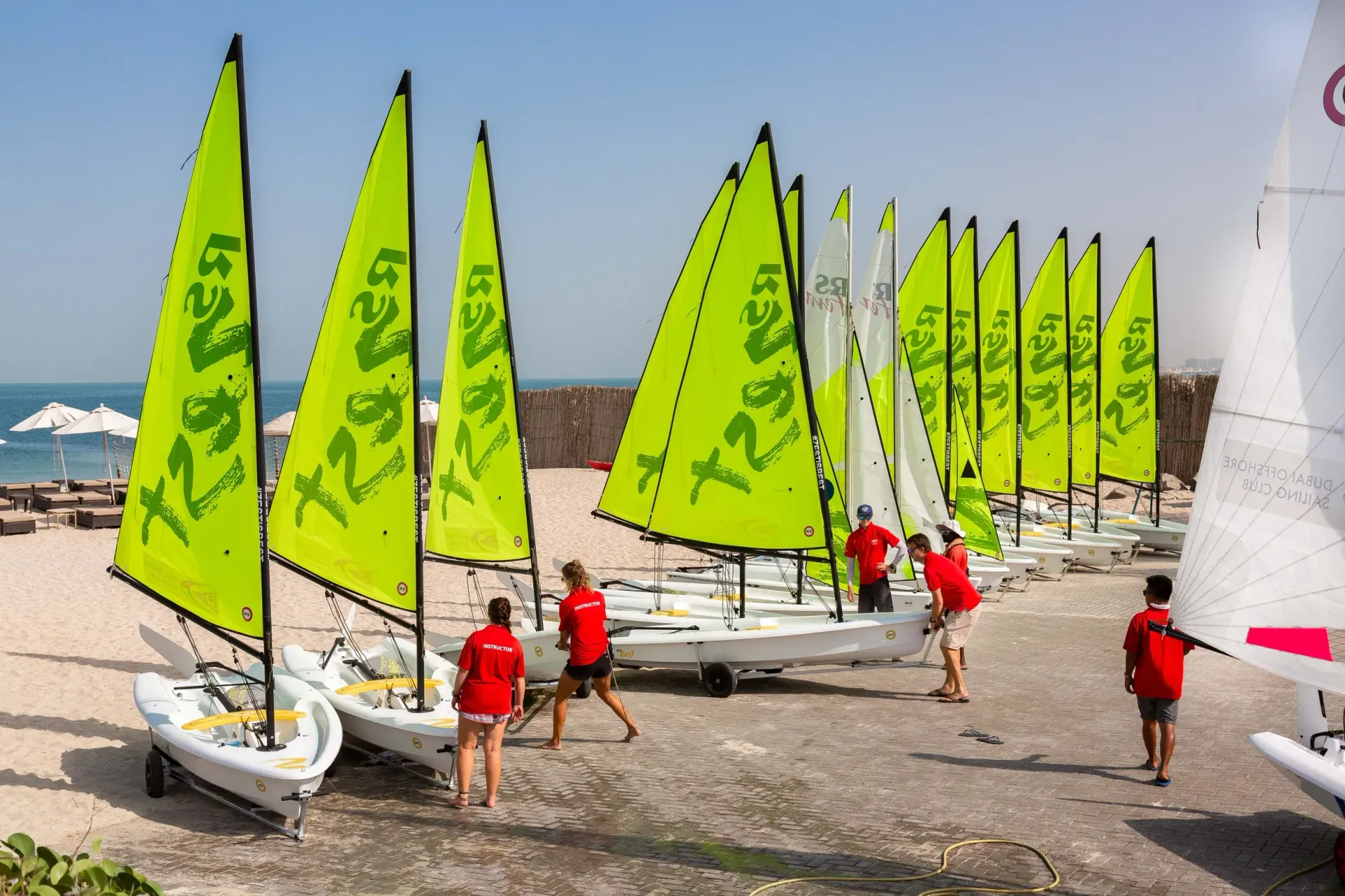 Bright lime-green sailboats lined up on a beach with several people in red life vests.