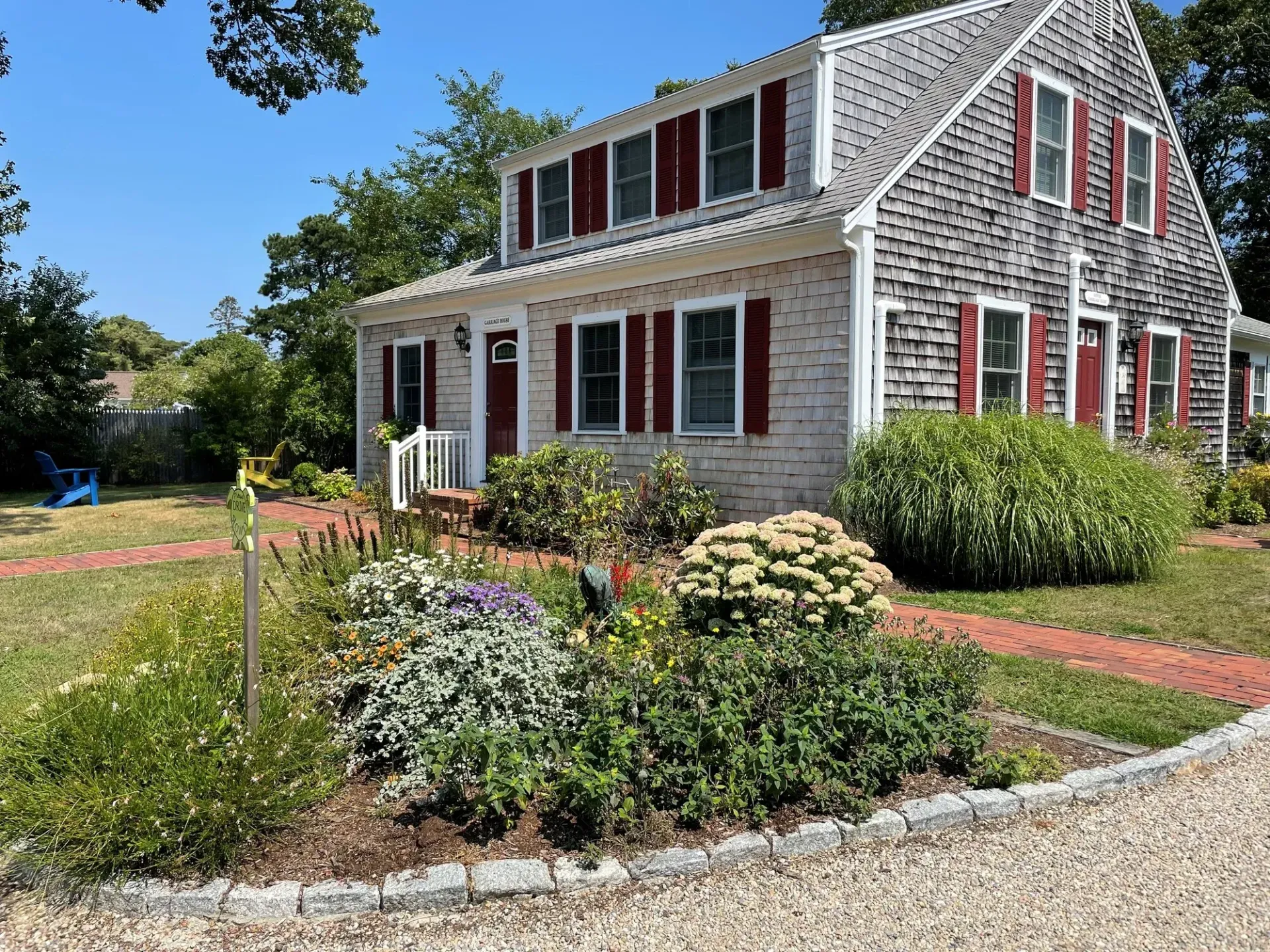 Gray shingled house with red shutters, a blooming front garden, and a brick walkway on a sunny day
