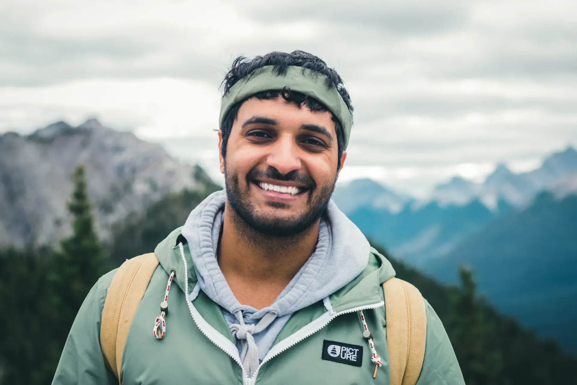 Smiling hiker in a green jacket and headband with mountains and cloudy sky in the background