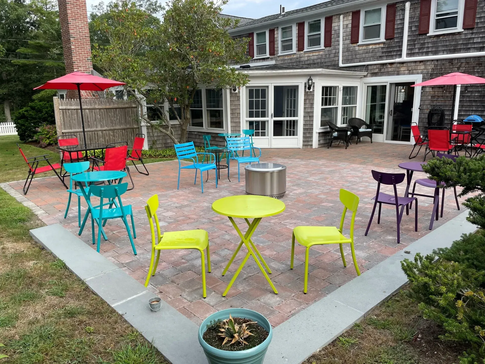 Colorful patio with metal tables and chairs outside a house, including red umbrellas and a brick paver courtyard