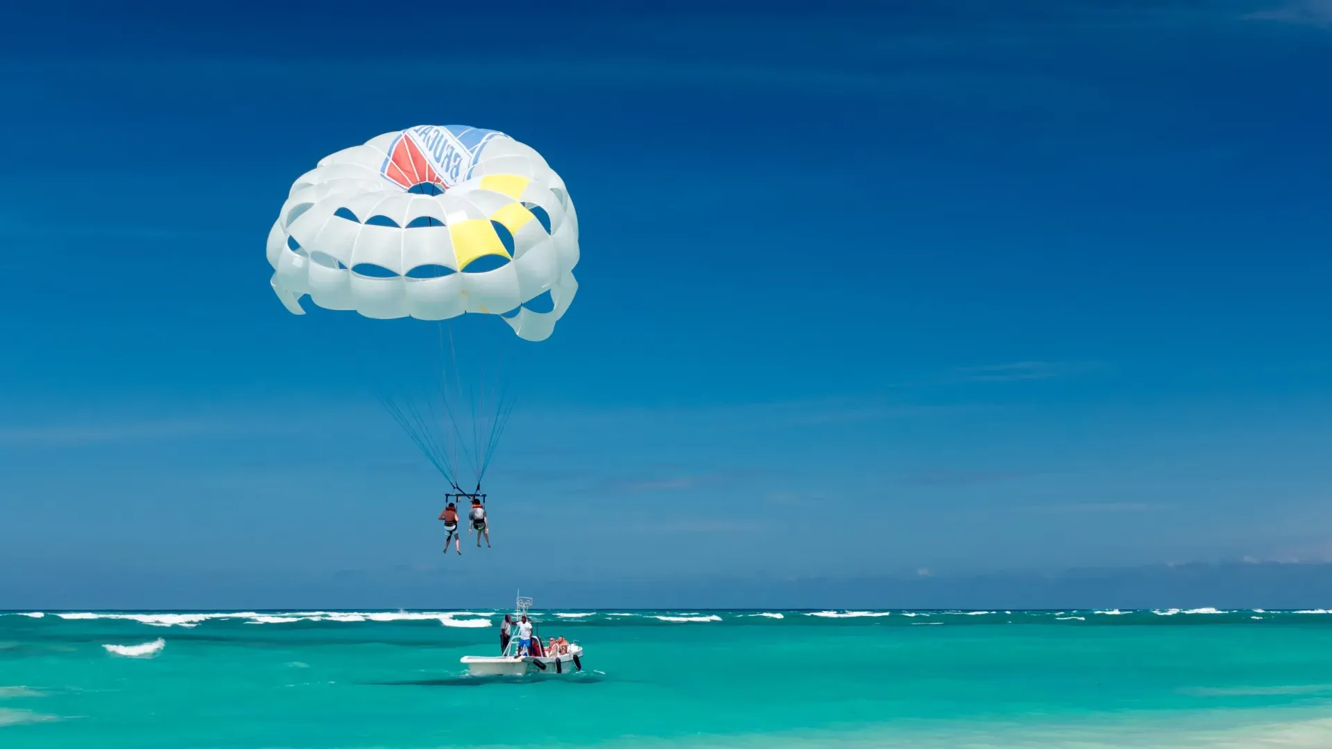 Parasailing over a turquoise sea with a white parachute under a clear blue sky.