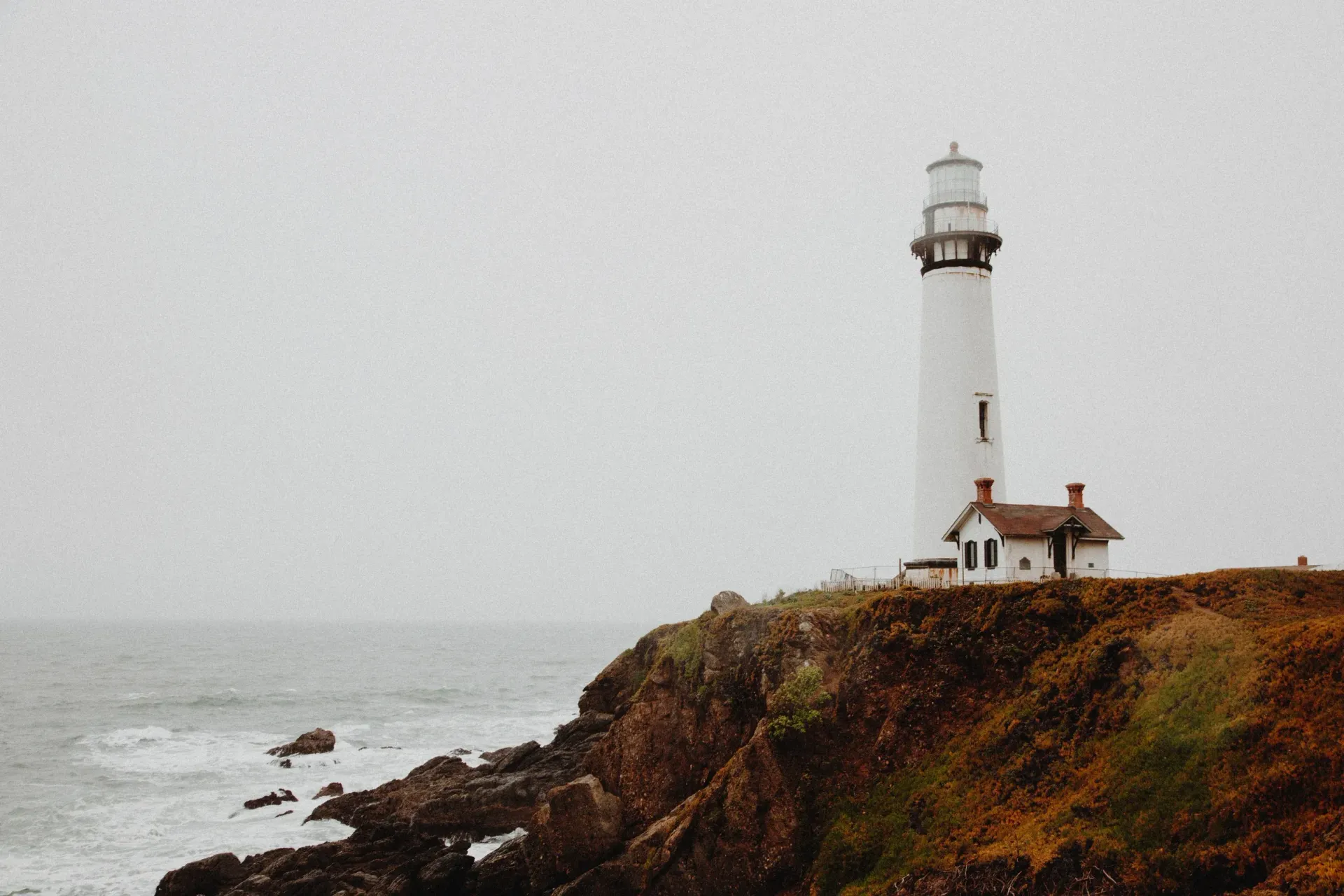 White lighthouse on a rocky seaside cliff under a gray, misty sky