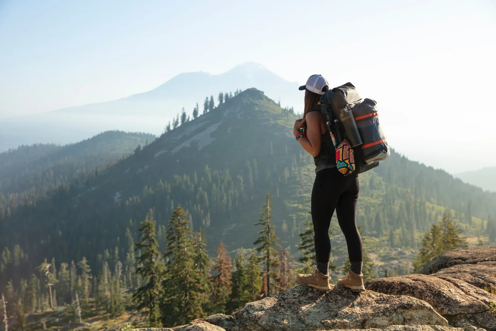 Hiker with backpack standing on a rocky ridge overlooking forested mountains at sunrise