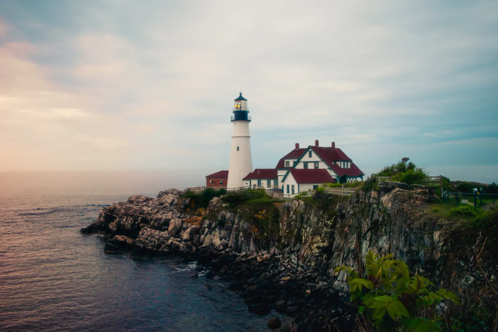 Lighthouse and red-roofed house on rocky seaside cliff at sunset