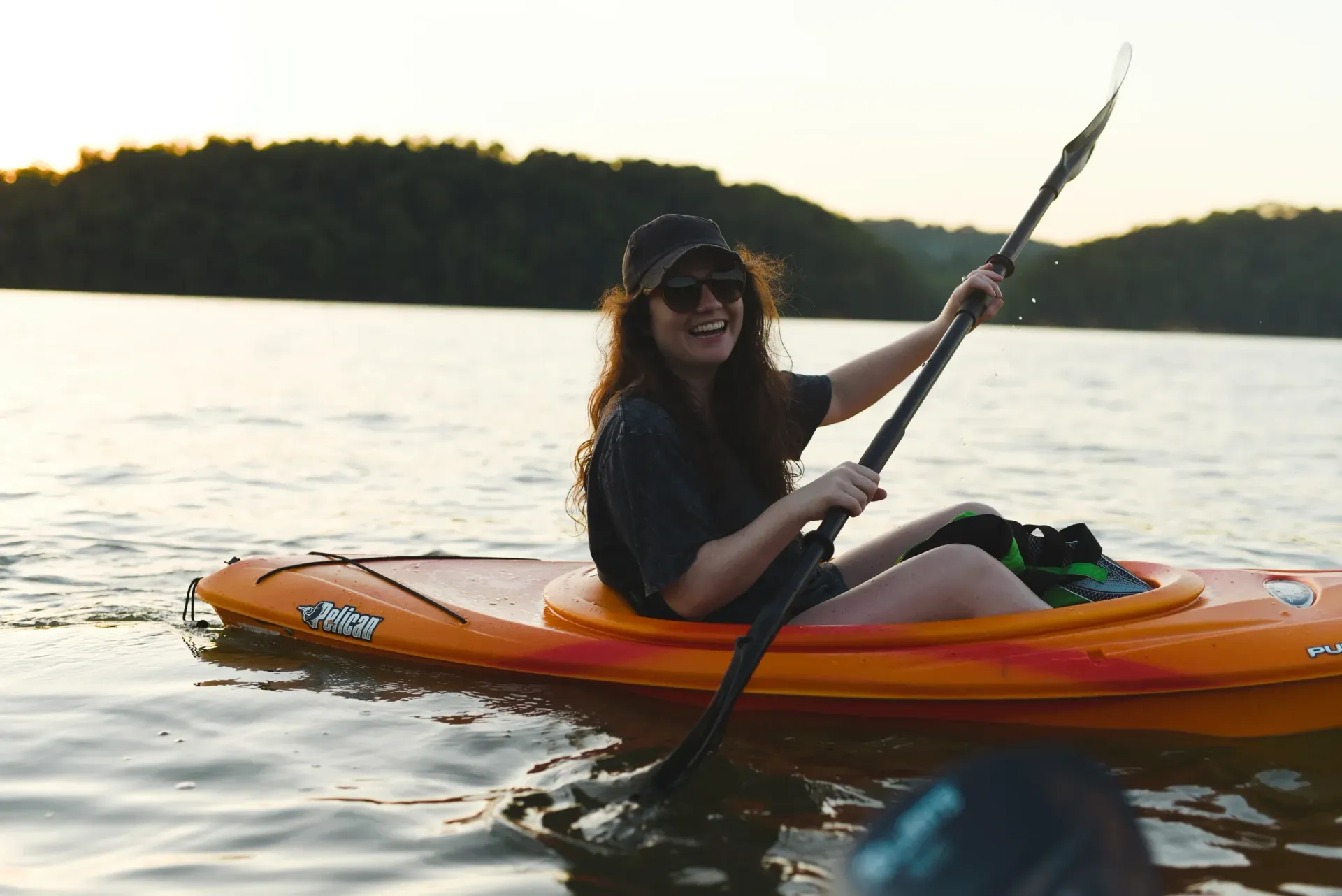 Person kayaking in an orange kayak on a calm lake at sunset, holding a paddle and smiling