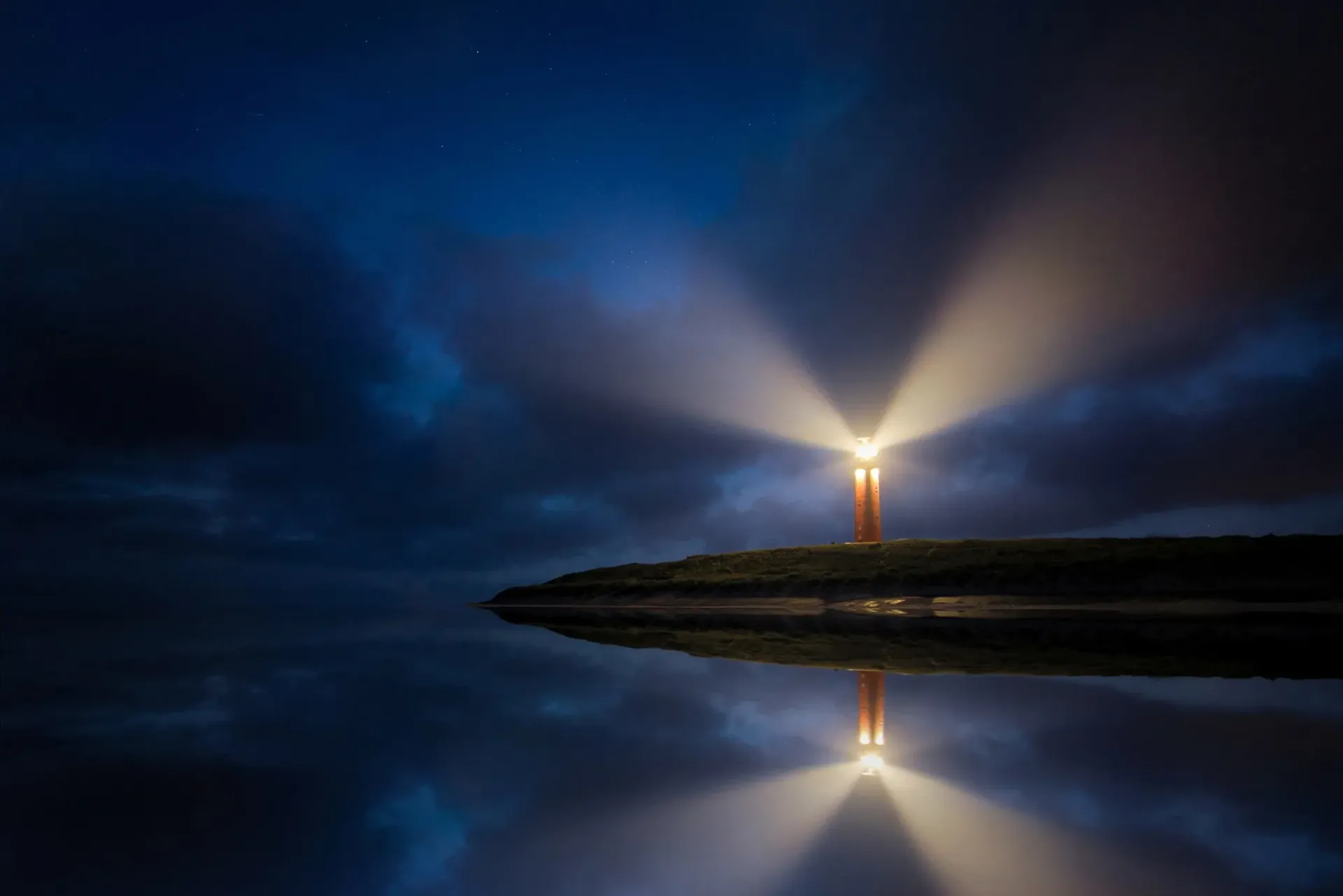 Lighthouse beam at night over calm water, with a bright reflection and dark blue sky.