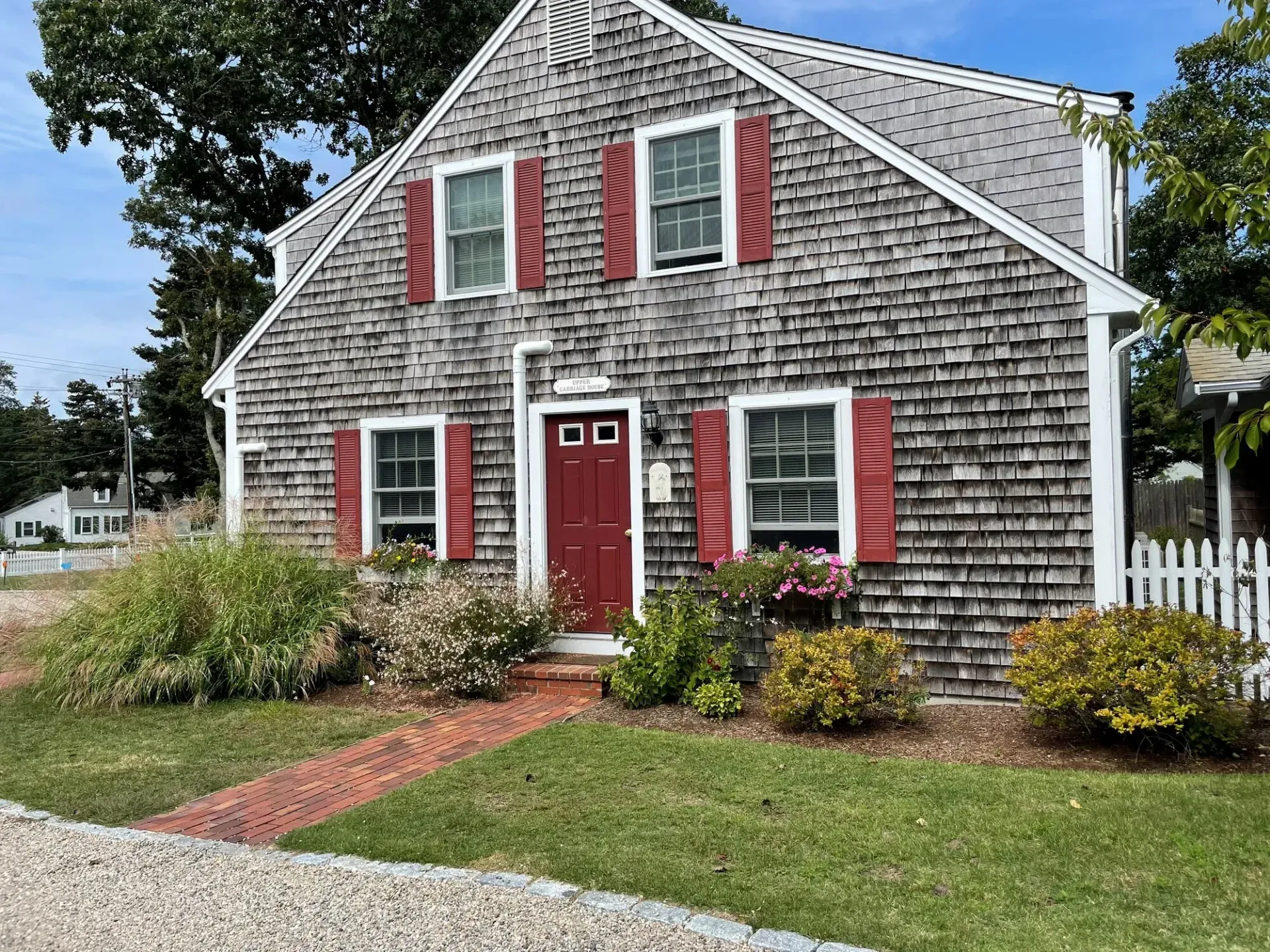 Red-shuttered gray shingle house with a red door, brick path, and white picket fence