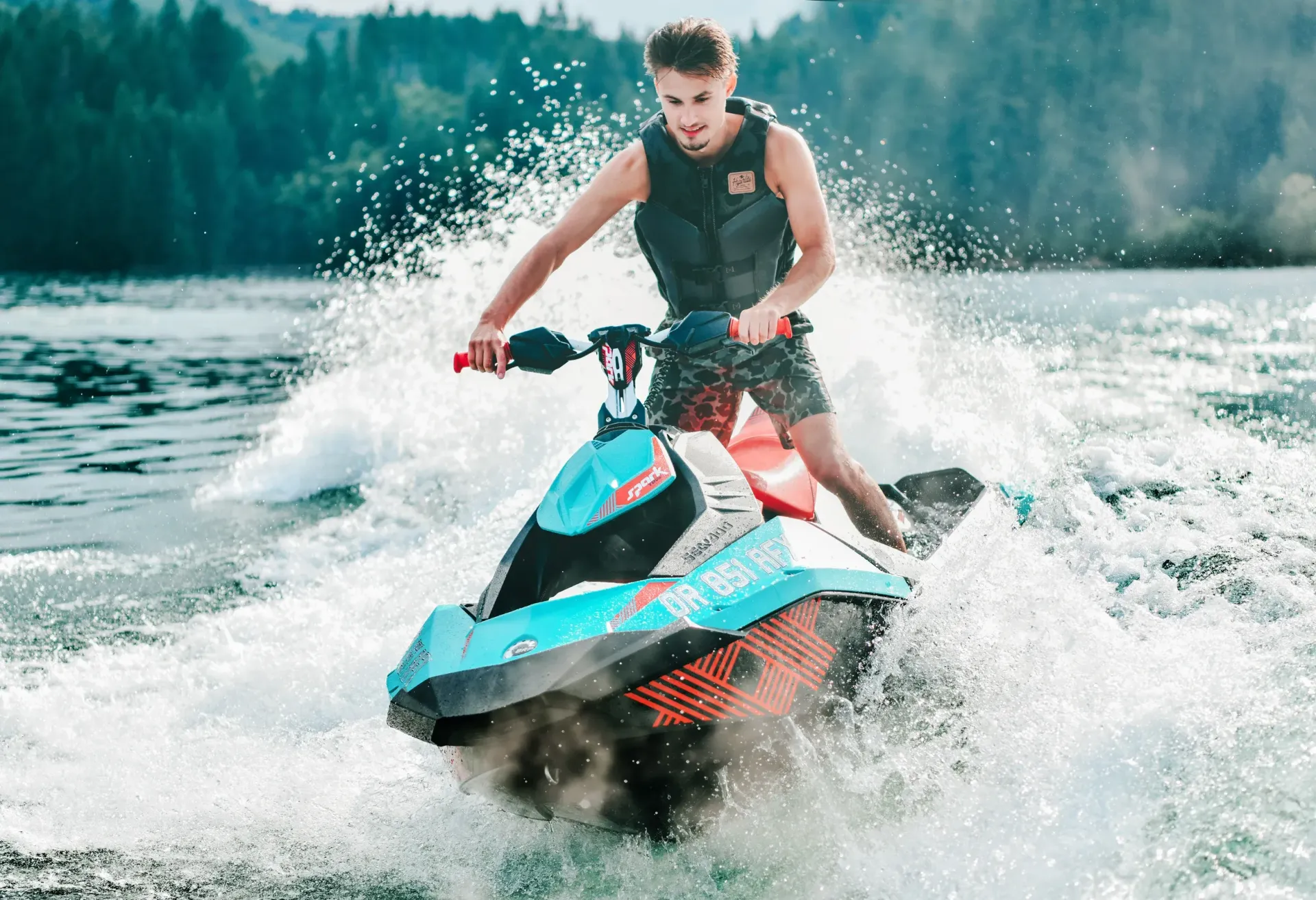 Person riding a teal and pink jet ski through splashing water on a lake with forested shore behind