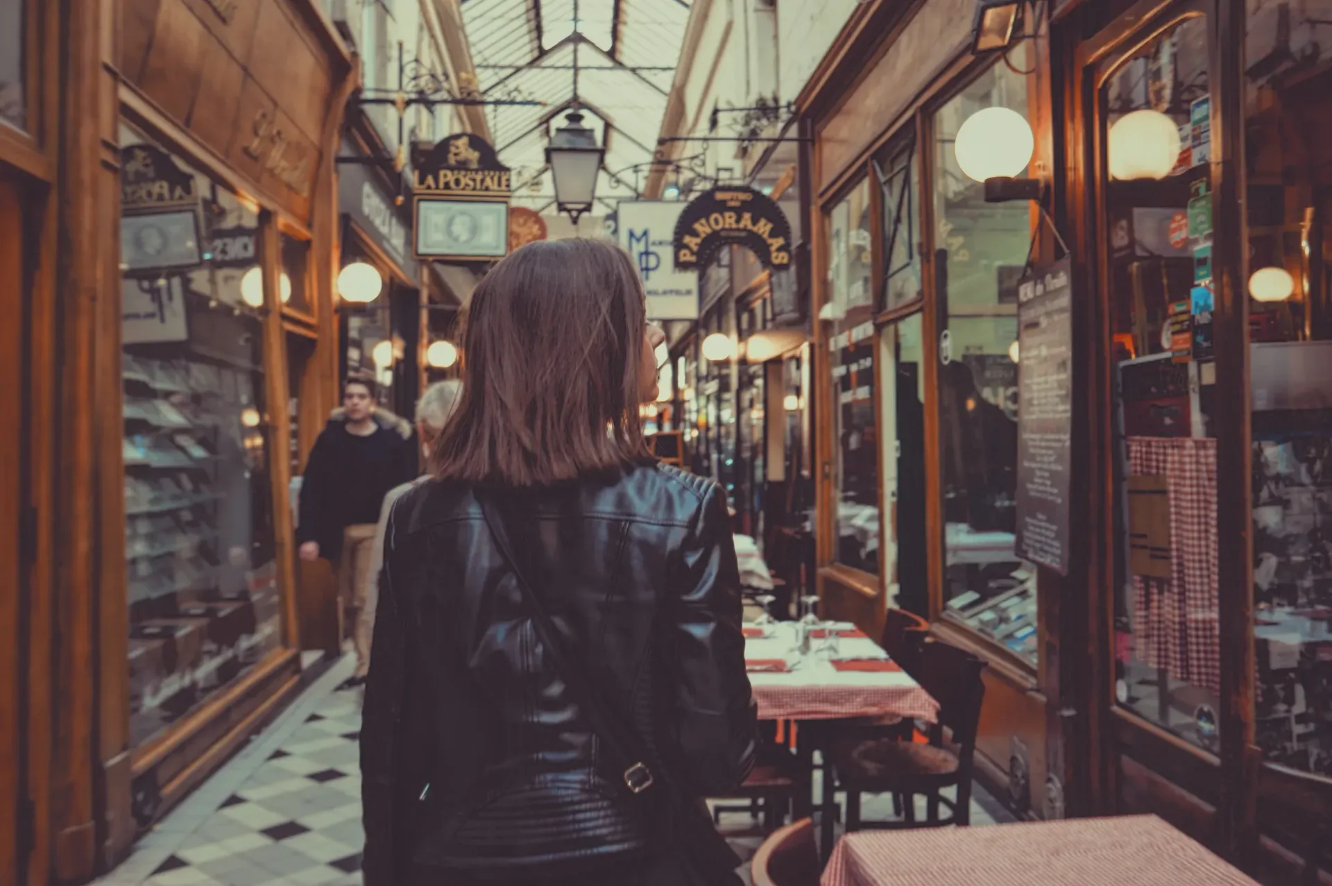 Woman walking through a narrow café-lined indoor arcade with warm lights and checkered floor