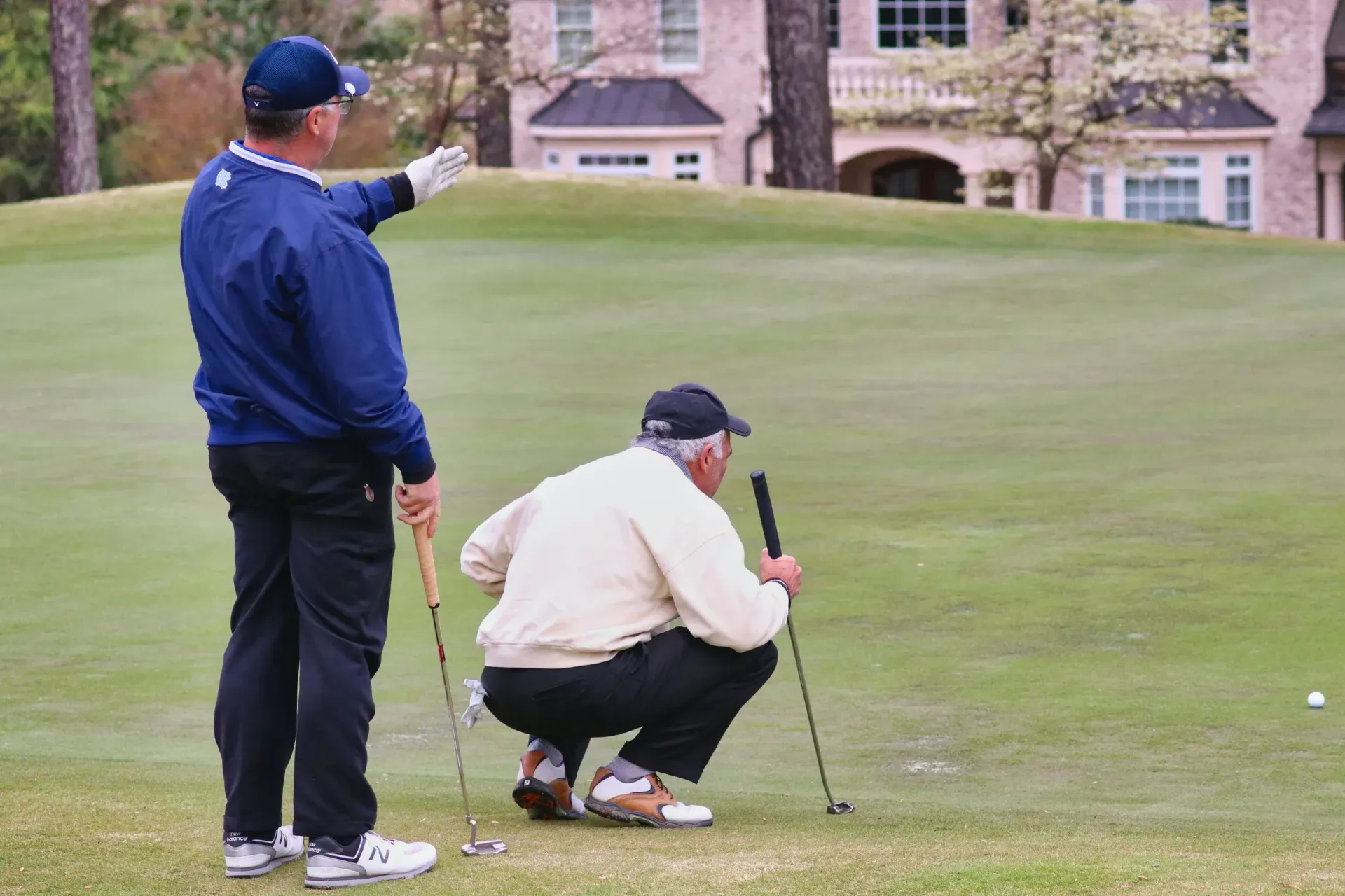 Two golfers on a green, one crouching to line up a putt while another points ahead.