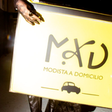 Persona sosteniendo un cartel amarillo brillante que dice “MDU MODISTA A DOMICILIO” con un pequeño icono de un coche.