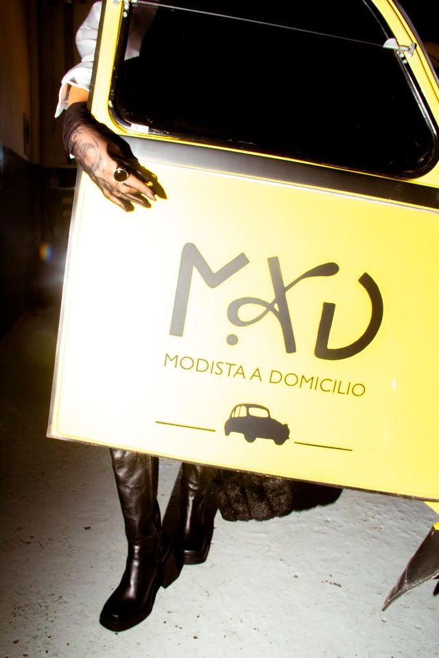 Persona sosteniendo un cartel amarillo de reparto con el logo de “Modista a Domicilio” y el icono de un coche.