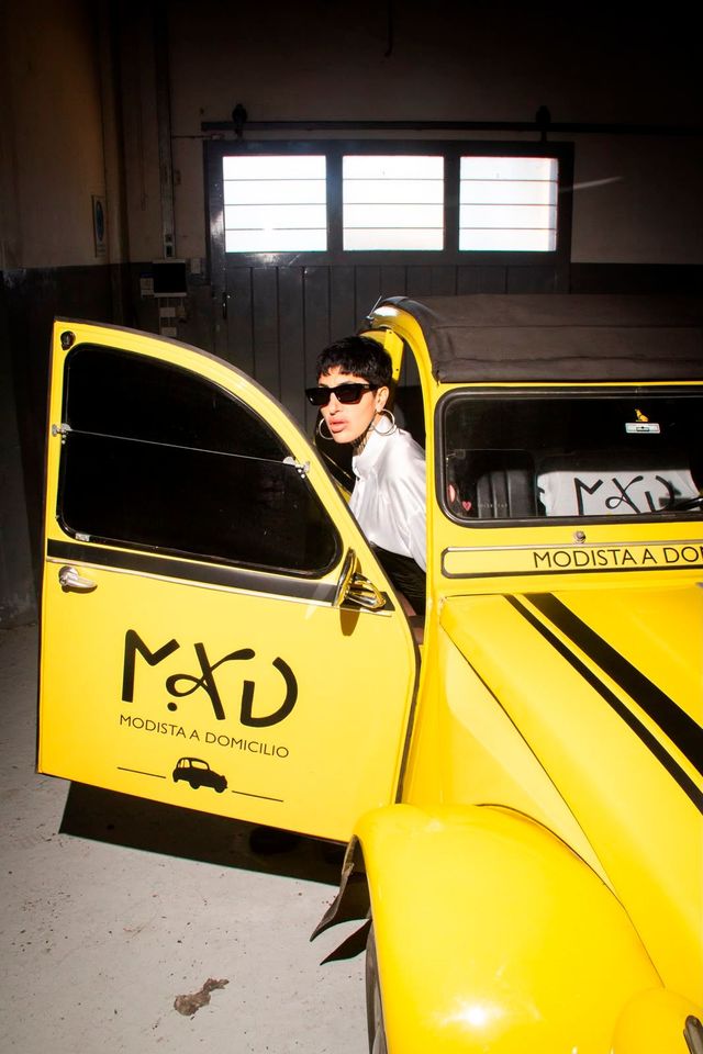 Una persona con gafas de sol se asoma por la ventanilla de un coche amarillo con la puerta abierta en un espacio que parece un garaje.