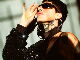 Mujer con gafas de sol y top negro posando con la mano cerca de la cara, luciendo grandes pendientes de aro y un tatuaje en el cuello.