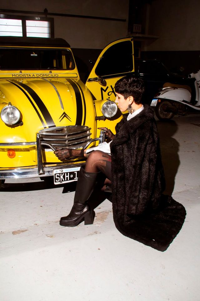 Persona agachada junto a un Volkswagen Beetle amarillo en un garaje, vestida con un abrigo de piel oscuro.