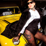 Persona vestida de negro y con gafas de sol posando sobre un coche antiguo amarillo.