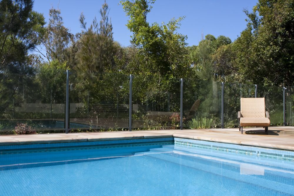 Blue Pool, Glass Fence, Trees, and Wooden Lounge Chair — East Coast Pool Resurfacing In Port Macquarie, NSW