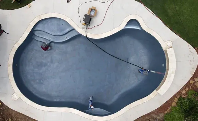 Aerial view of a dark blue, kidney-shaped swimming pool with people working inside; gray concrete surround. — East Coast Pool Resurfacing In Port Macquarie, NSW