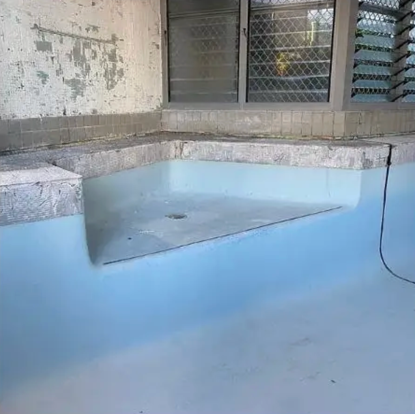 A light blue swimming pool corner bench submerged in water, set against a weathered wall and a window with a metal grate. — East Coast Pool Resurfacing In Port Macquarie, NSW
