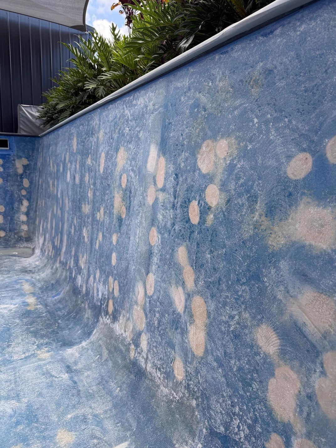 An empty swimming pool wall with blue textured coating and faded white patches, viewed at an angle outdoors.  — East Coast Pool Resurfacing In Port Macquarie, NSW