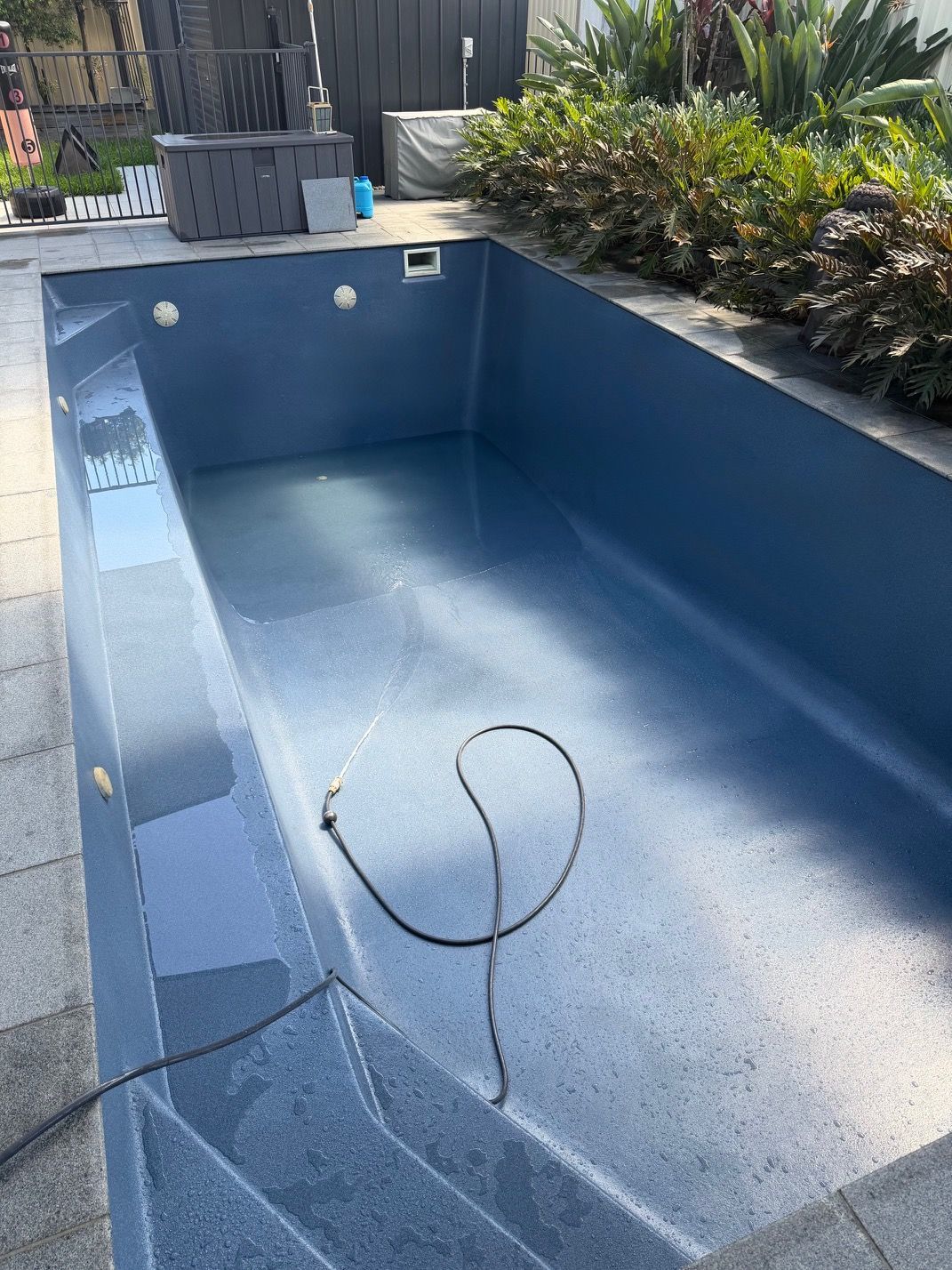 An empty, rectangular backyard swimming pool with dark blue, speckled interior walls and built-in steps. — East Coast Pool Resurfacing In Port Macquarie, NSW