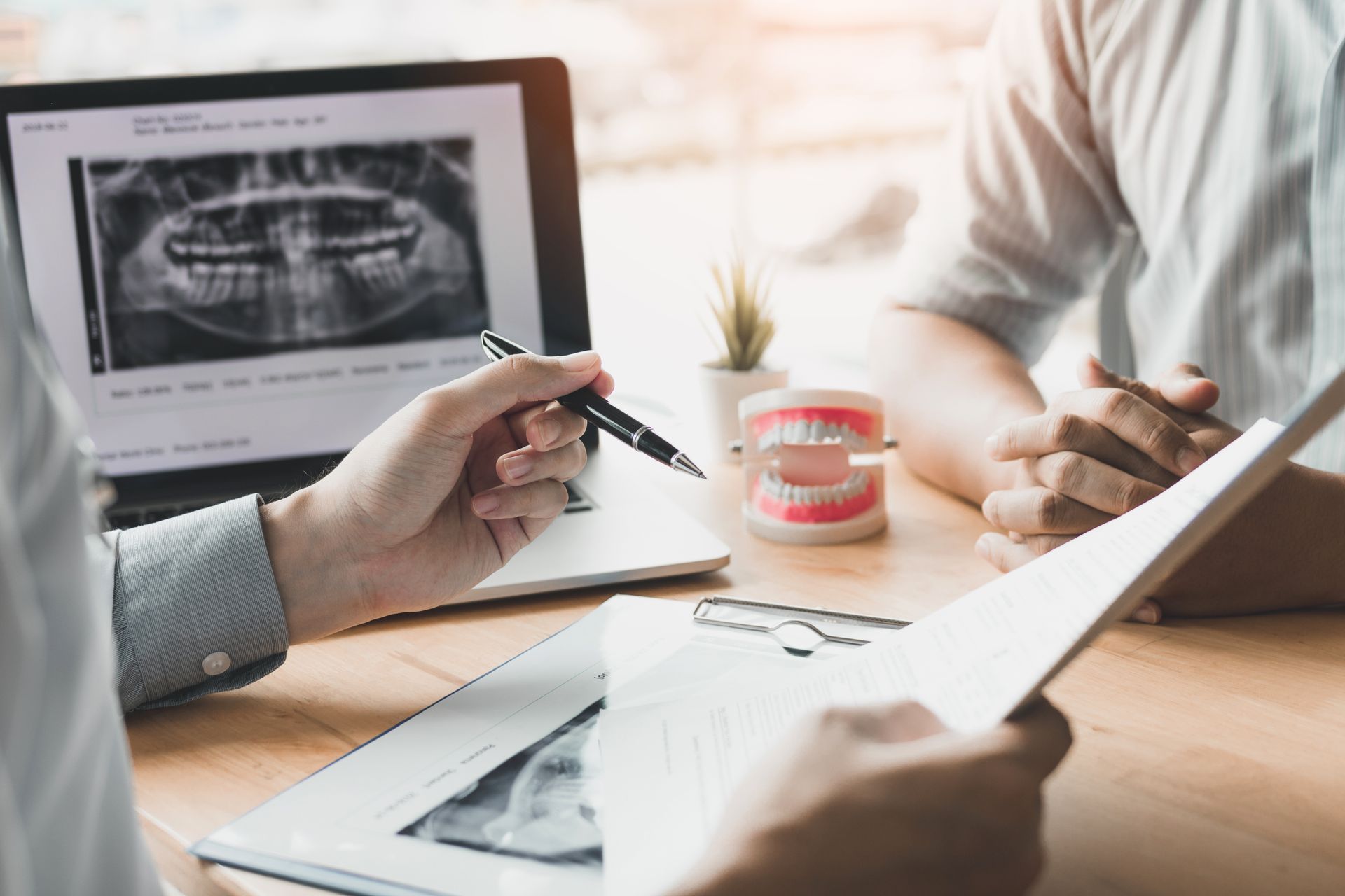 A dentist uses a pen to explain a digital dental X-ray and a model of teeth to a patient during a consultation.