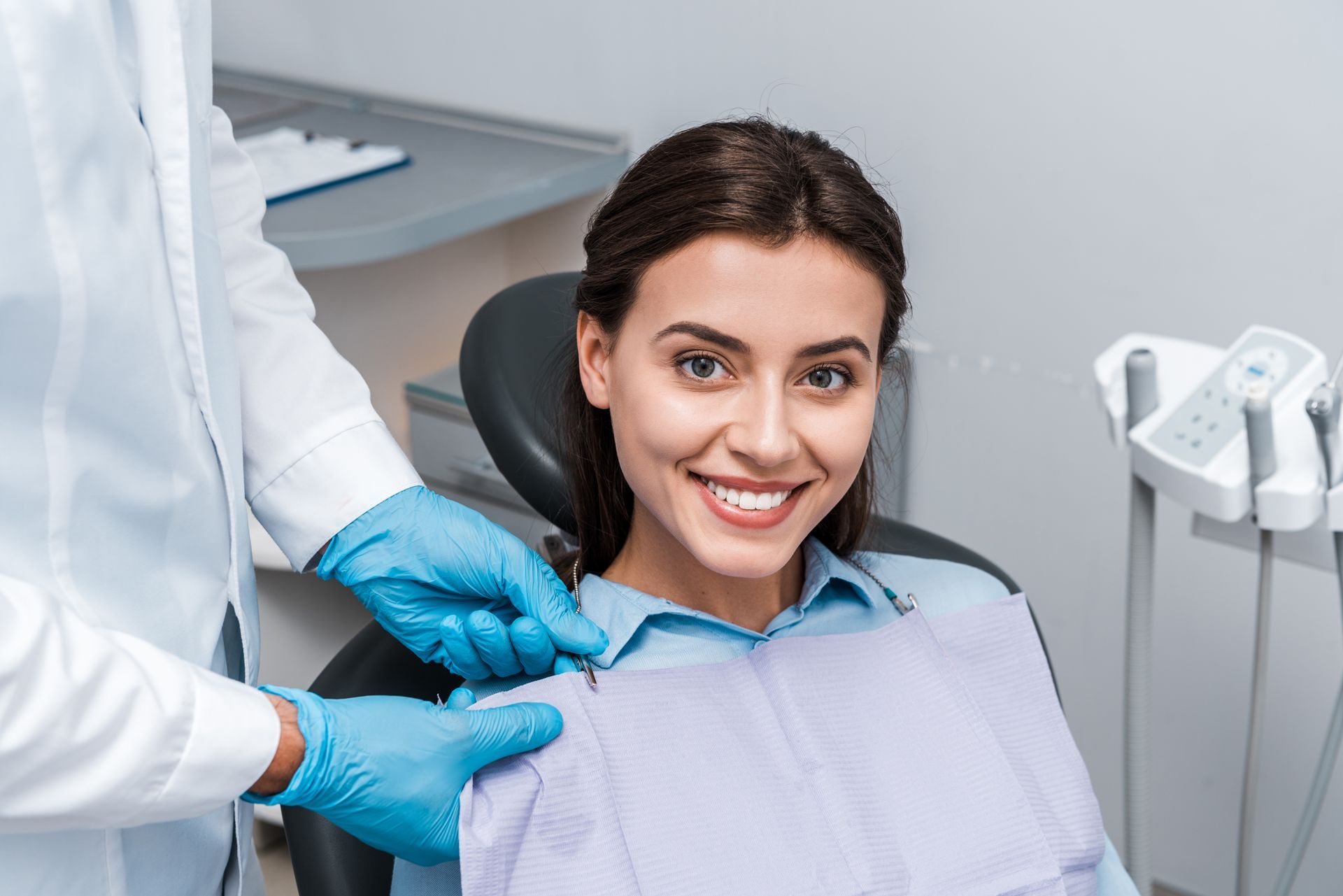 A dentist in a green uniform and blue gloves examines a smiling patient’s teeth in a clinical setting.